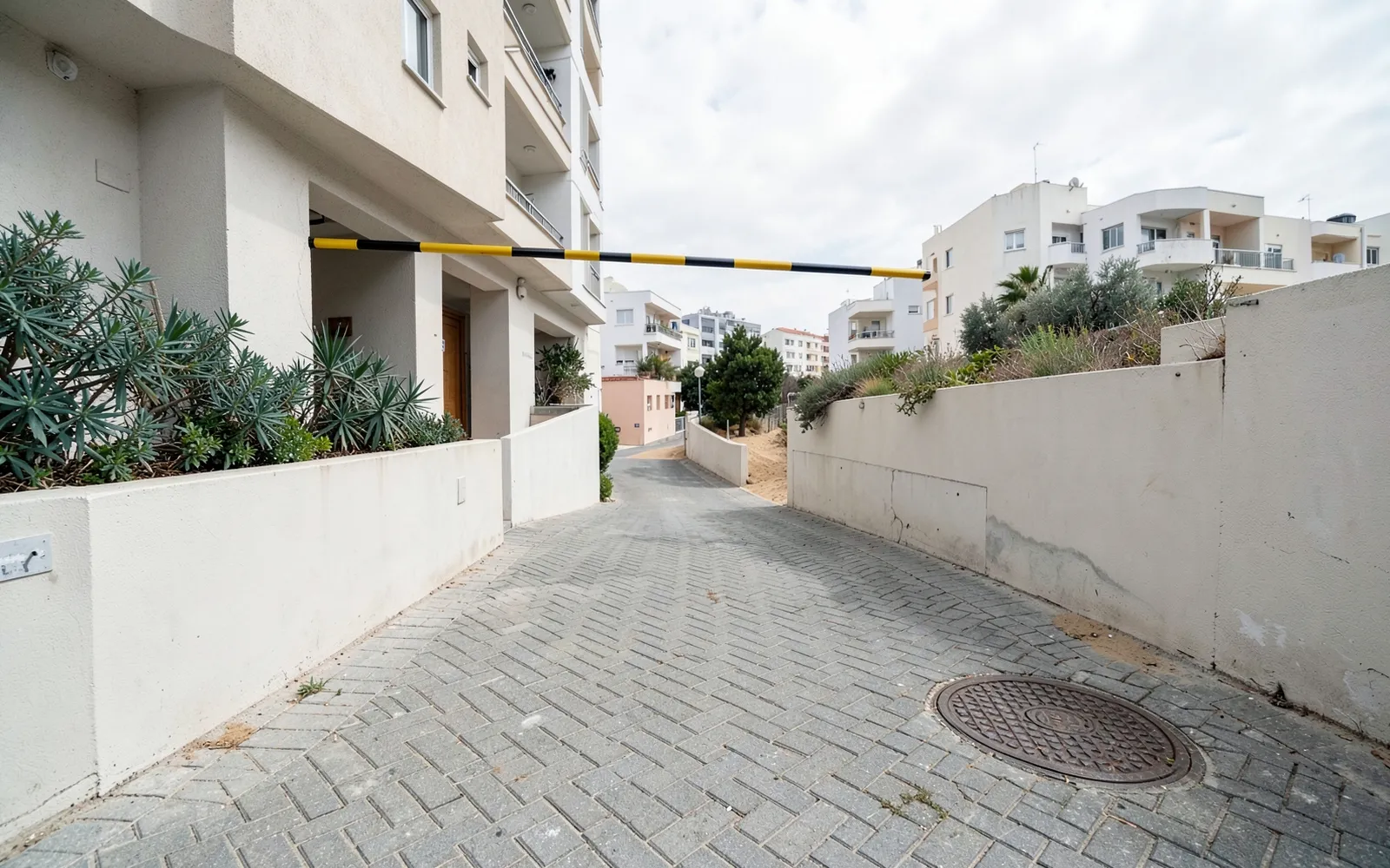 מעבר מרוצף אל חניון של בניין מגורים, עם מוט הגבלת גובה צהוב-שחור מעל הכניסה ומכסה ביוב עגול בריצוף